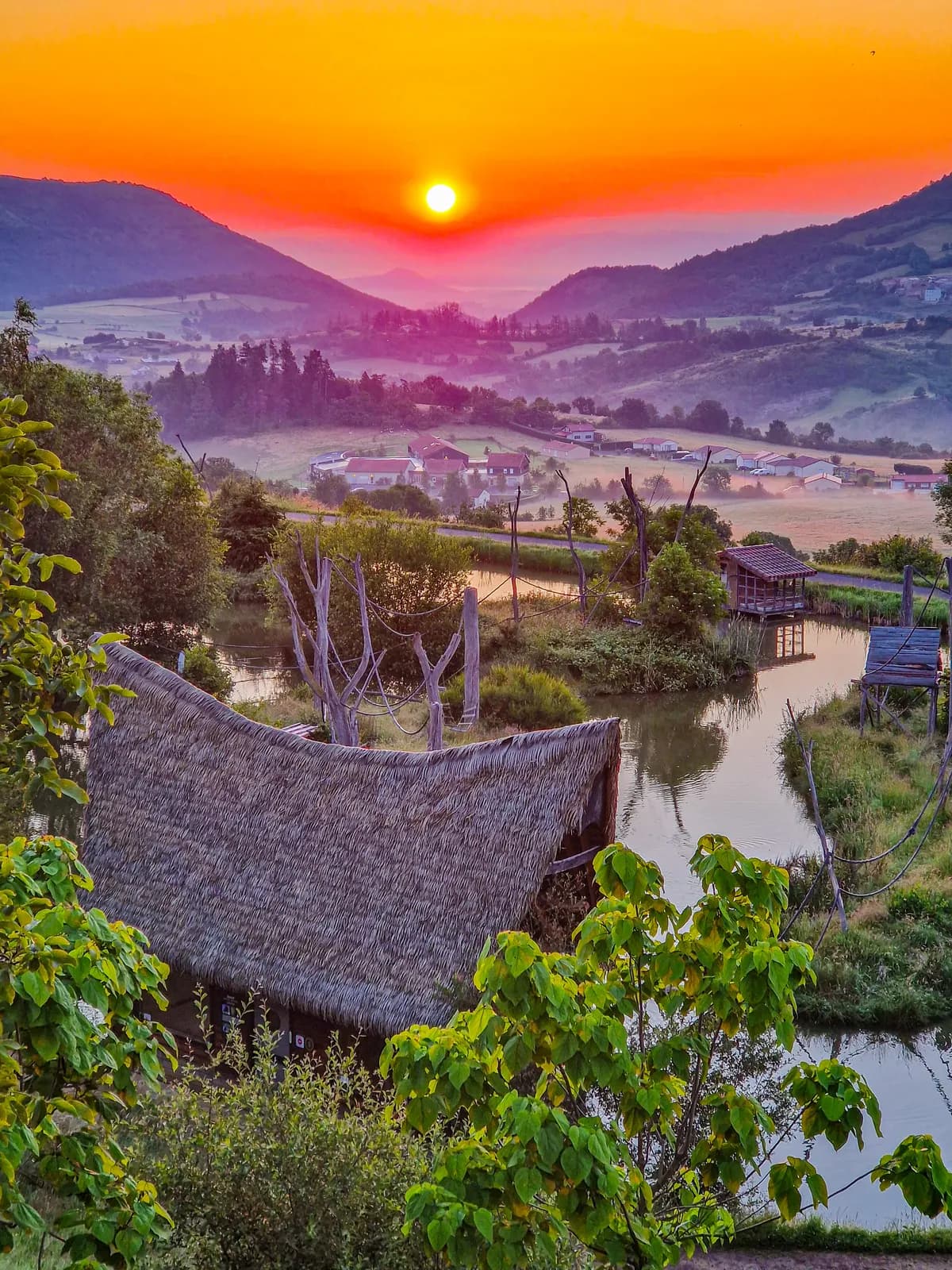 Le soleil se lève sur les monts d’Auvergne : un toit de chaume au premier plan, un étang, et un village au creux de la vallée.